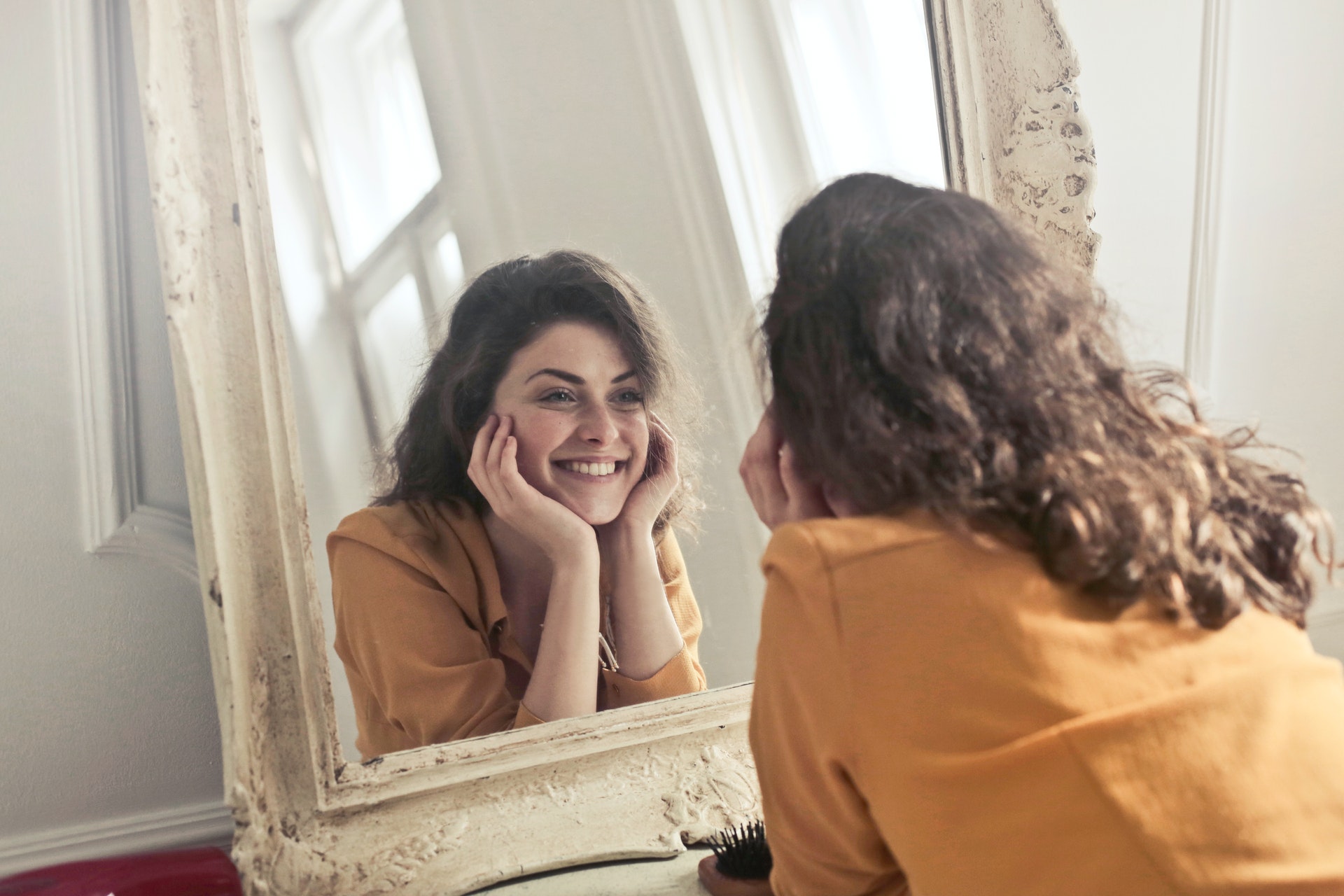 a woman is looking at herself in the mirror. She is smiling and her hands are cupping her face.