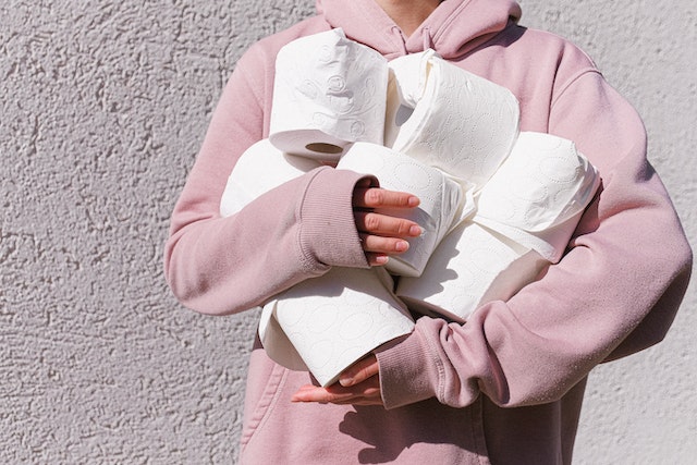 A person in a pink sweatshirt holding 7 rolls of toilet paper in their arms