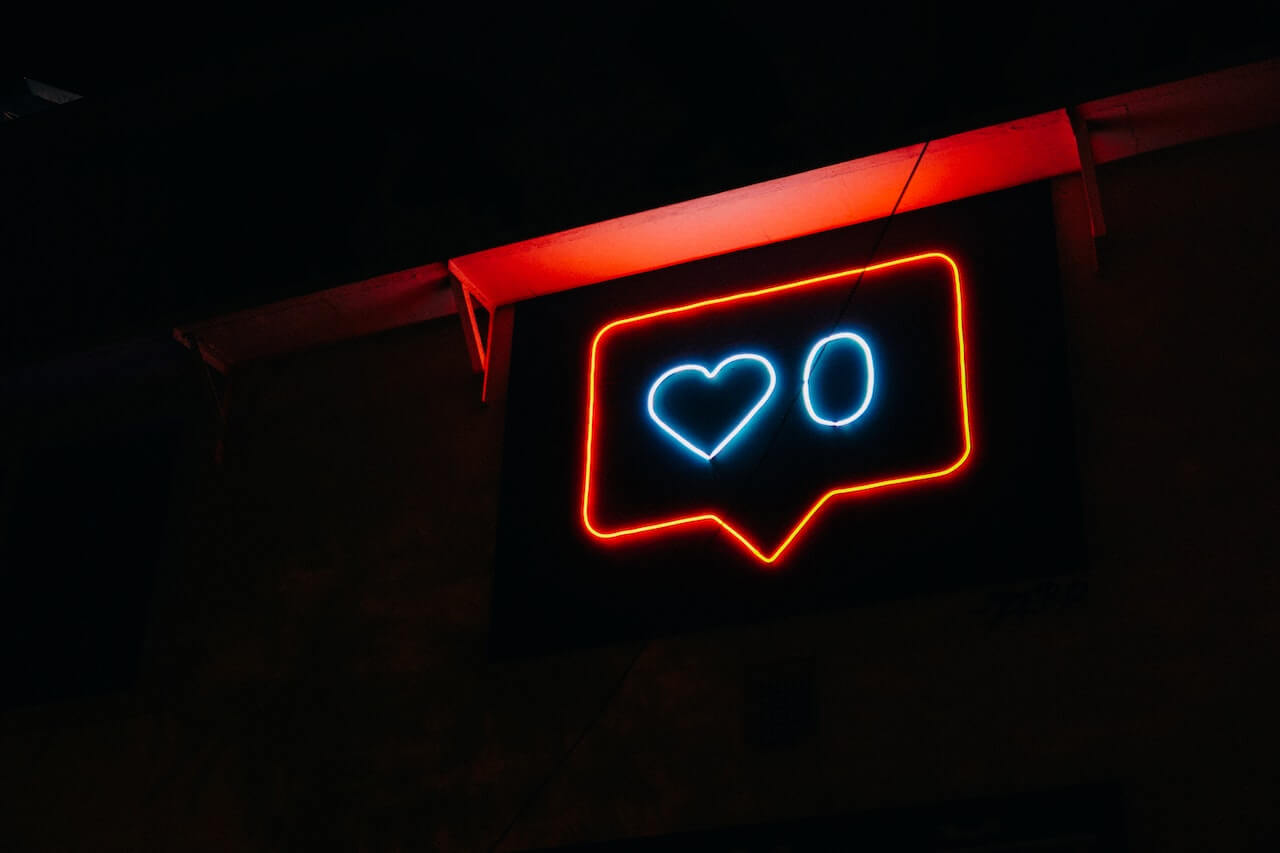 A glowing neon sign of a red speech bubble in the dark. Inside the bubble is the blueish-white outline of a heart and a zero next to it, representing zero likes.