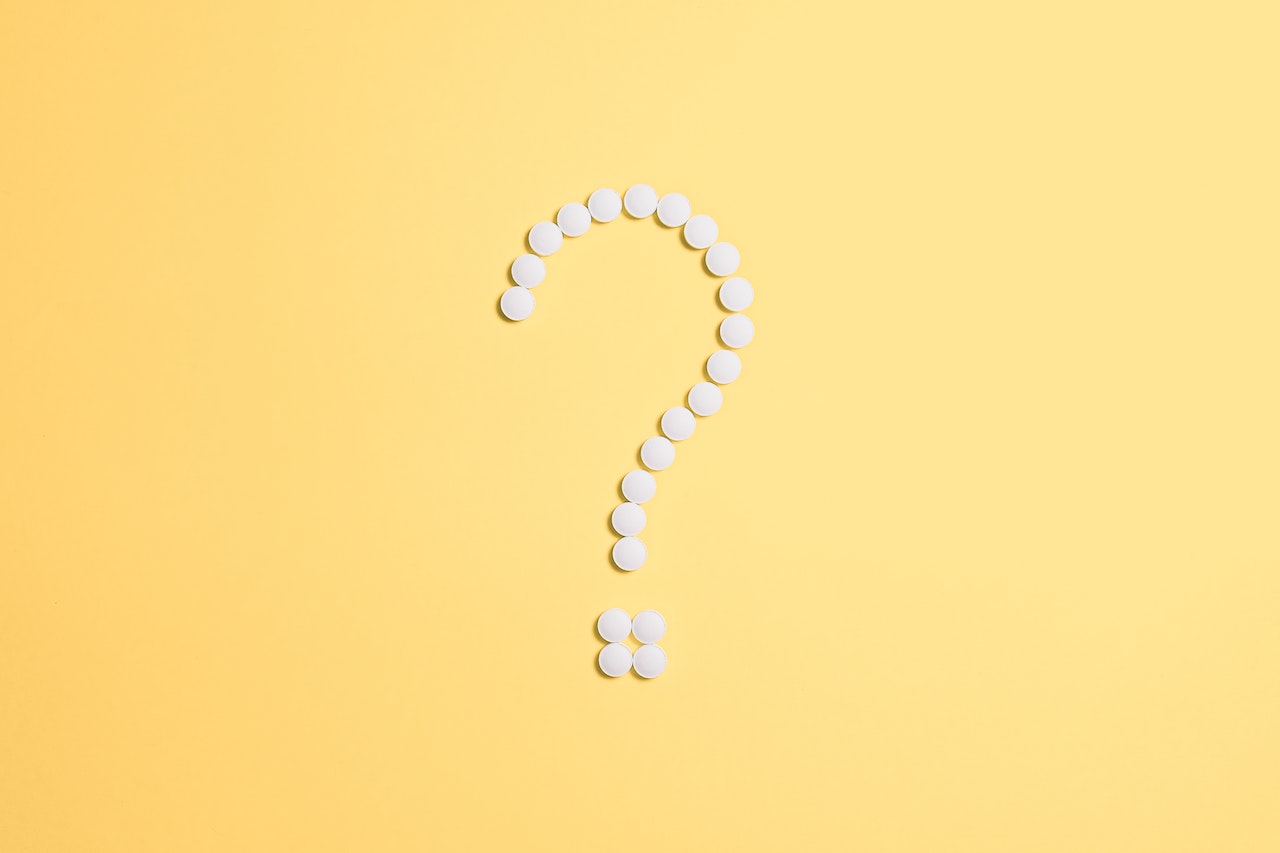 Several white pills on a yellow background, arranged into the shape of a question mark.