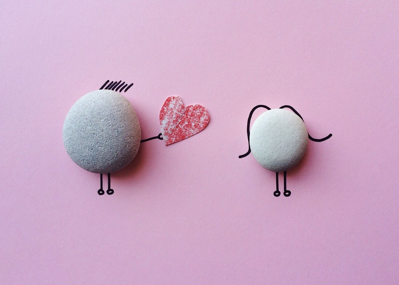 two rocks on a pink background. One rock is handing a heart to the other rock. Photo by Pixabay.