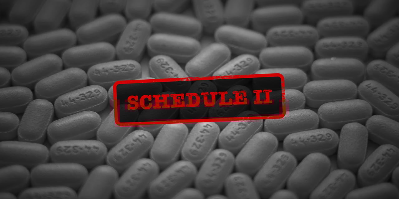 black and white photo of a variety of pills on a surface. Over the pills in bright red is a stamp that reads, "schedule ii"