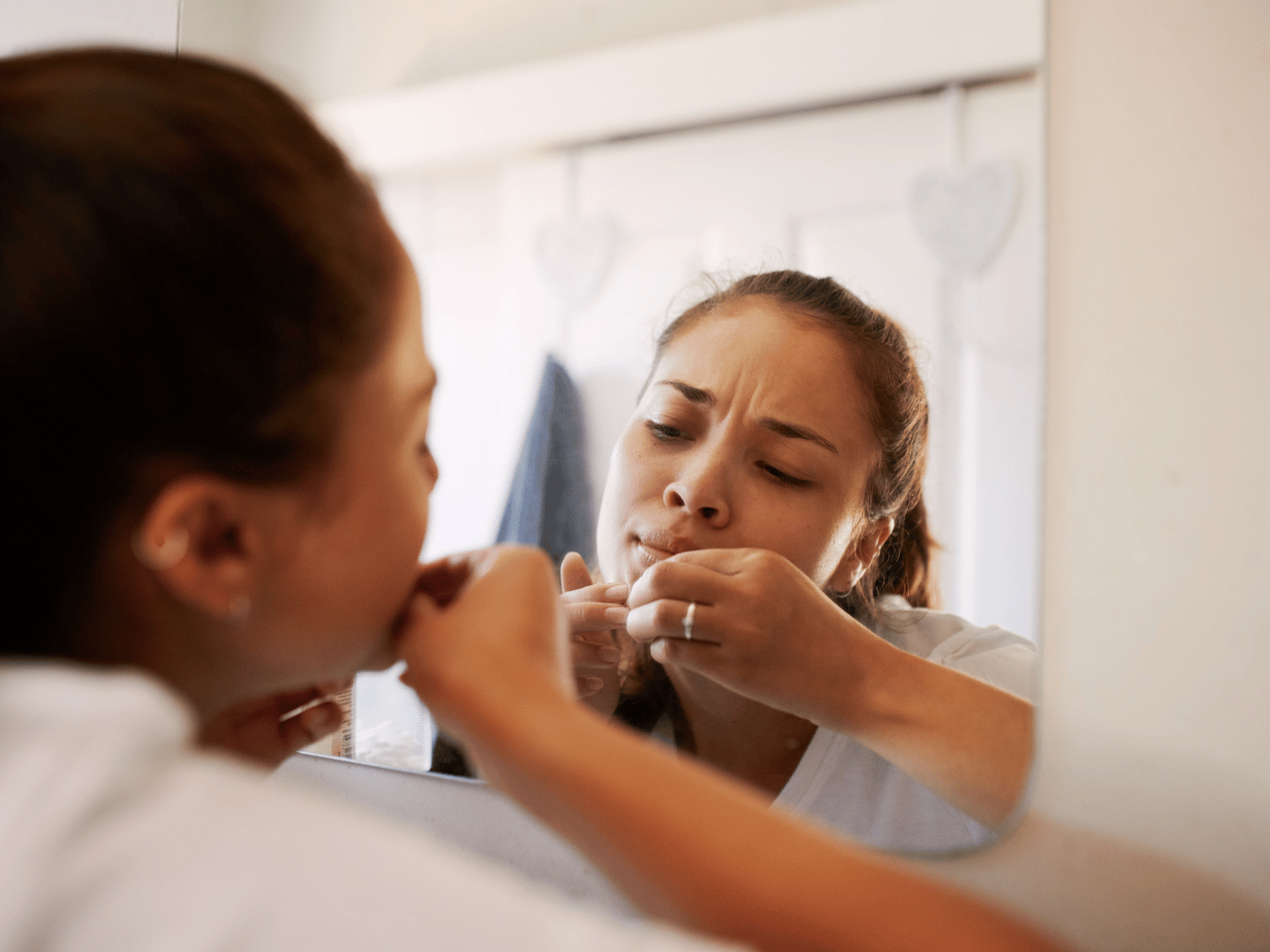 woman leaning into her reflection in the mirror to get a better look at the blemishes on her chin that she's picking at.