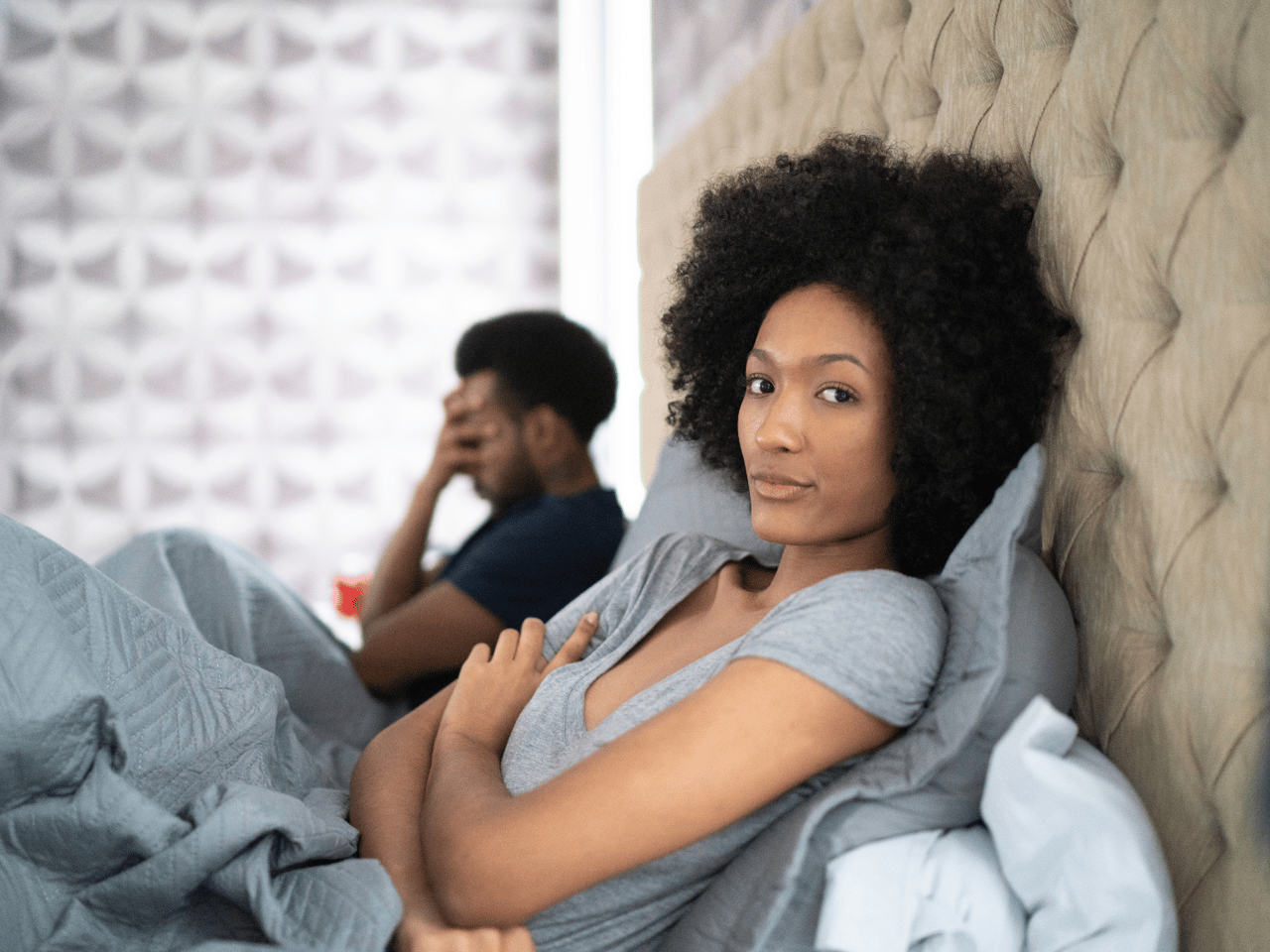A Black couple in bed together. The man is in the background with his hand covering her face, and the woman is in the foreground, looking at the camera with a bored expression.