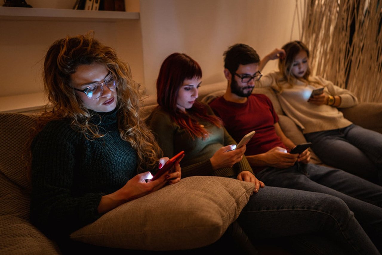 Four young adults sit on a sofa in a dimly lit room looking at their illuminated phone screen.