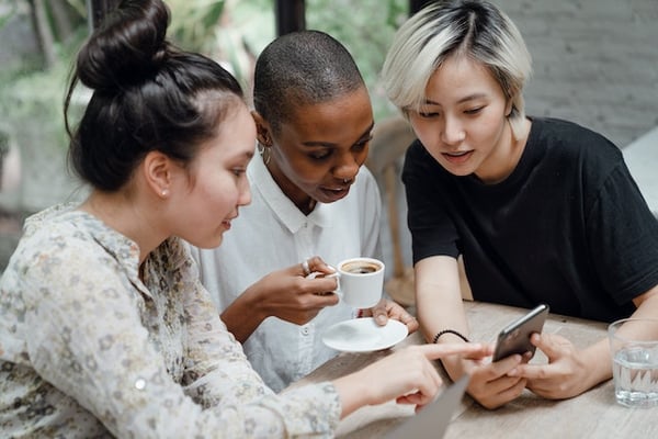 People with ADHD tend to be forgetful, impulsive, and/or inattentive, which can complicate how well we adhere to the etiquette of texting. Image shows three friends sitting around a table looking at a single phone while one person explains how to adjust settings for text messages.