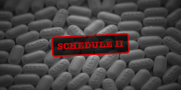 black and white photo of a variety of pills on a surface. Over the pills in bright red is a stamp that reads, "schedule ii"