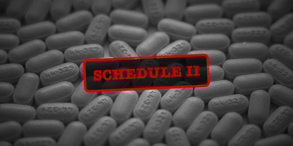 black and white photo of a variety of pills on a surface. Over the pills in bright red is a stamp that reads, "schedule ii"