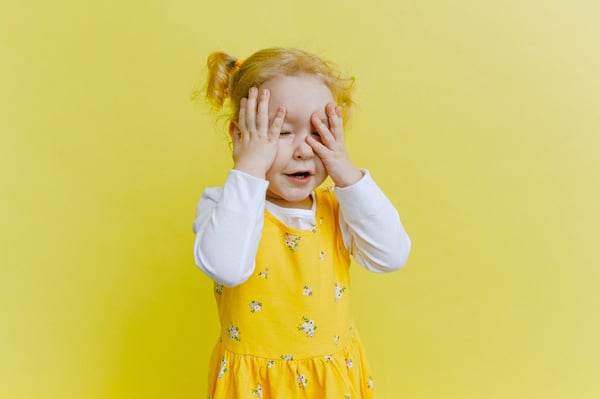 Bright yellow background with a girl, toddler-aged, hiding her face. photo by Anna Shvets.