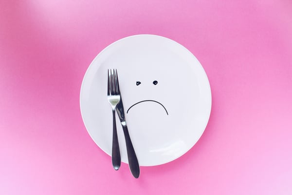 a white plate with only a fork and knife on it. there is a sad face drawn on the plate, and the background is pink.