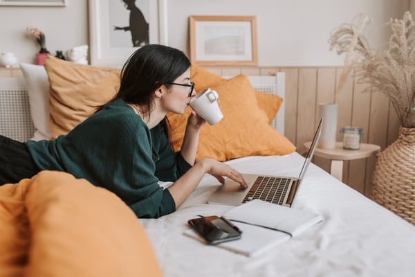 woman laying on a bed, drinking coffee, and on her laptop. She is surrounded by her phone and notebooks.