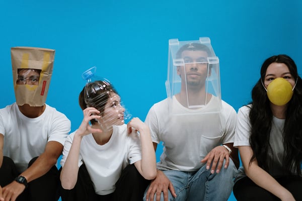 Four young adults sitting in front of a blue wall. They each have a random object over their head or face, like a plastic bin and a paper bag. Photo by cottonbro from Pexels.