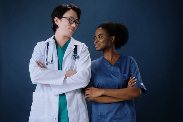 two doctors standing next to and smiling at each other with their arms crossed. One is an asian man and another is a black woman. photo by cottonbro on pexels.