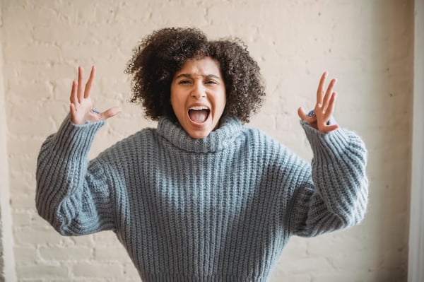 Woman in blue knit sweater, her arms raised in an angry gesture, is yelling at the camera.