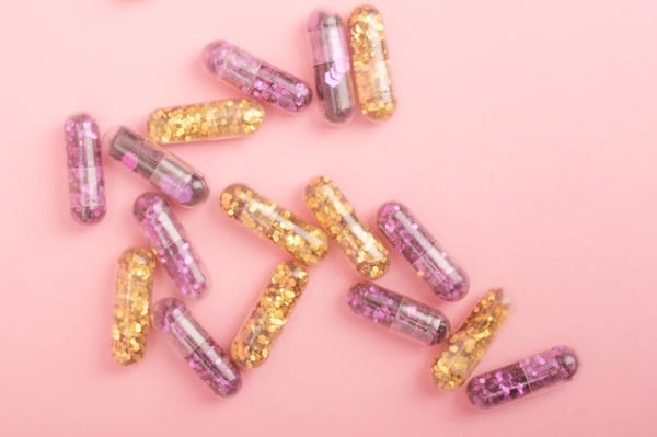 A pink background with pill capsules containing two different colors of glitter to symbolize adderall and vyvanse comparison for adhd medication.