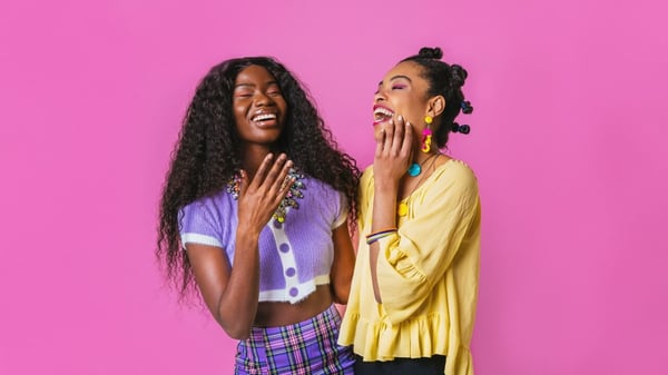 Two Black women with ADHD standing in front of a bright pink background are laughing and smiling together