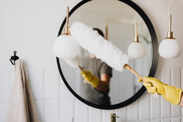 The image shows a section of a bathroom, large round bathroom mirror, 2 white lamps, one of which is being dusted with a large feather duster. The mirror reflection shows the person who is cleaning. The face is obscured and the reflection is blurry.