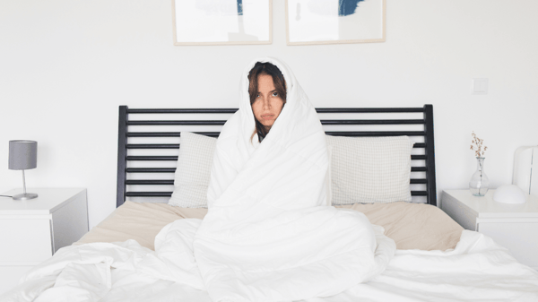 A woman with ADHD sitting up in bed in the morning with the blankets over her head; she's tired and wants to stay in bed and go back to sleep.