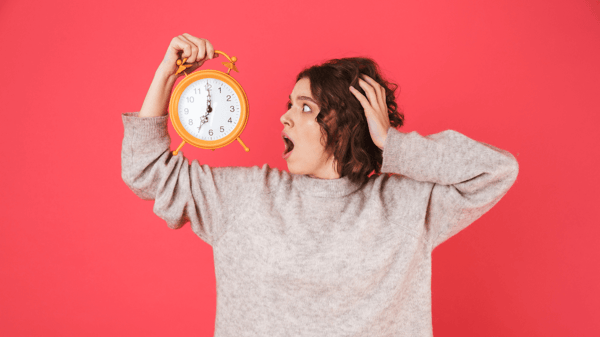 A woman holding up an alarm clock and looking at it with stressed expression on her face