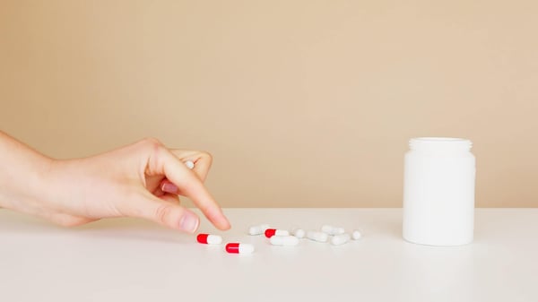 Loose white and red-and-white pills next to a white pill container, with a hand picking up one of the pills.