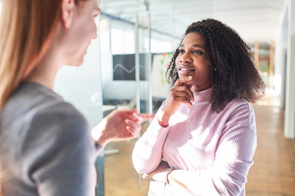 A white person wearing a grey top with long red hair in the image foreground is slightly out of focus. They are gesturing with their hand, speaking to a black woman in a pink mock-neck sweater standing in front of them. The black woman rests her hand on her chin as she listens. 