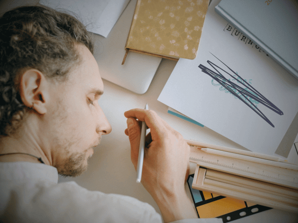 A person with light brown curly hair and patchy facial hair is lying on the side of their face, eyes closed, a pen in their right hand, on a desk. They’re surrounded by a laptop, notebooks and notepads, sticky notes, a ruler, and colored pencils. In the top-right corner is a partially visible notepad with the word BURNOUT stamped on it. Underneath the stamped word, in turquoise handlettering, the words - Be creative - have been scribbled out with dark grey marker.