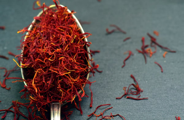 A closeup of a silver spoon, heaped with bright red saffron threads, on a dark grey background. Several saffron strands are scattered around the spoon. 