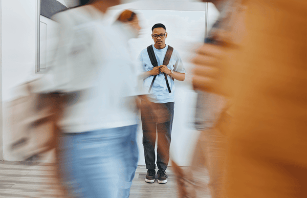 A man in glasses and a backpack stands in a white hallway facing the camera. People in the foreground walk quickly in front of him, giving a motion blur effect. 