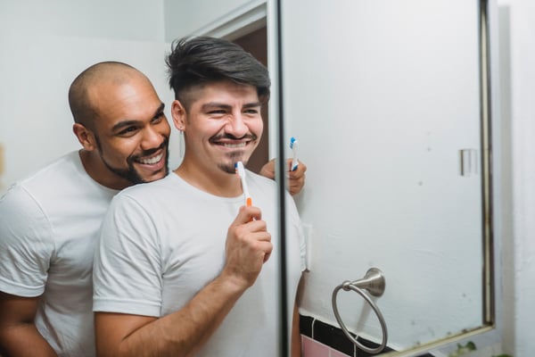 A couple - two men - brush their teeth together in the bathroom while smiling in the mirror. Photo by Ketut Subiyanto.