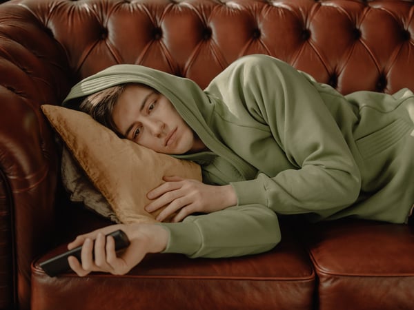 a young man is lying on a couch, looking bored. His hood is over his head and the remote is dangling from his hand.