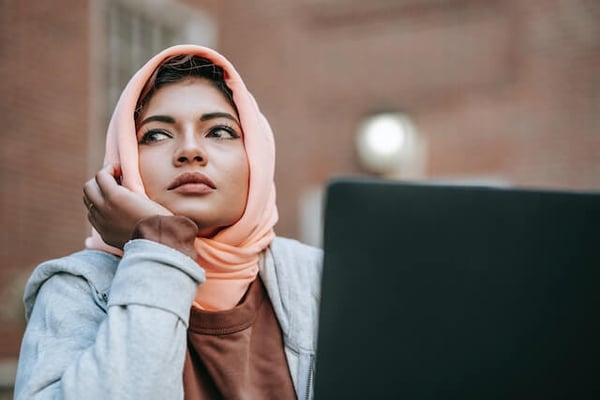 A woman with a head scarf is sitting at her laptop and staring off into space