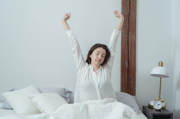 A light-skinned Bipoc woman in white pyjamas sitting up in bed and stretching her arms over her head. The room is quite bare, white walls, the bedsheets are white. There's an old-fashioned analog alarm clock on the bedside table.