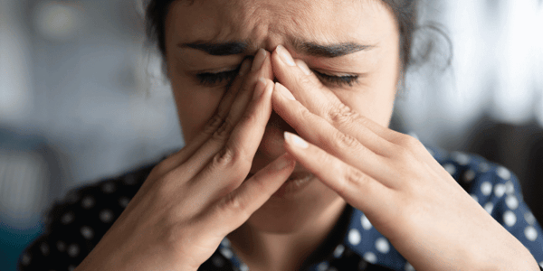 A young woman with brown skin and dark hair pinches the bridge of her nose with both hands due to stress at work. Her eyes are shut tight and she's on the verge of an emotional breakdown.