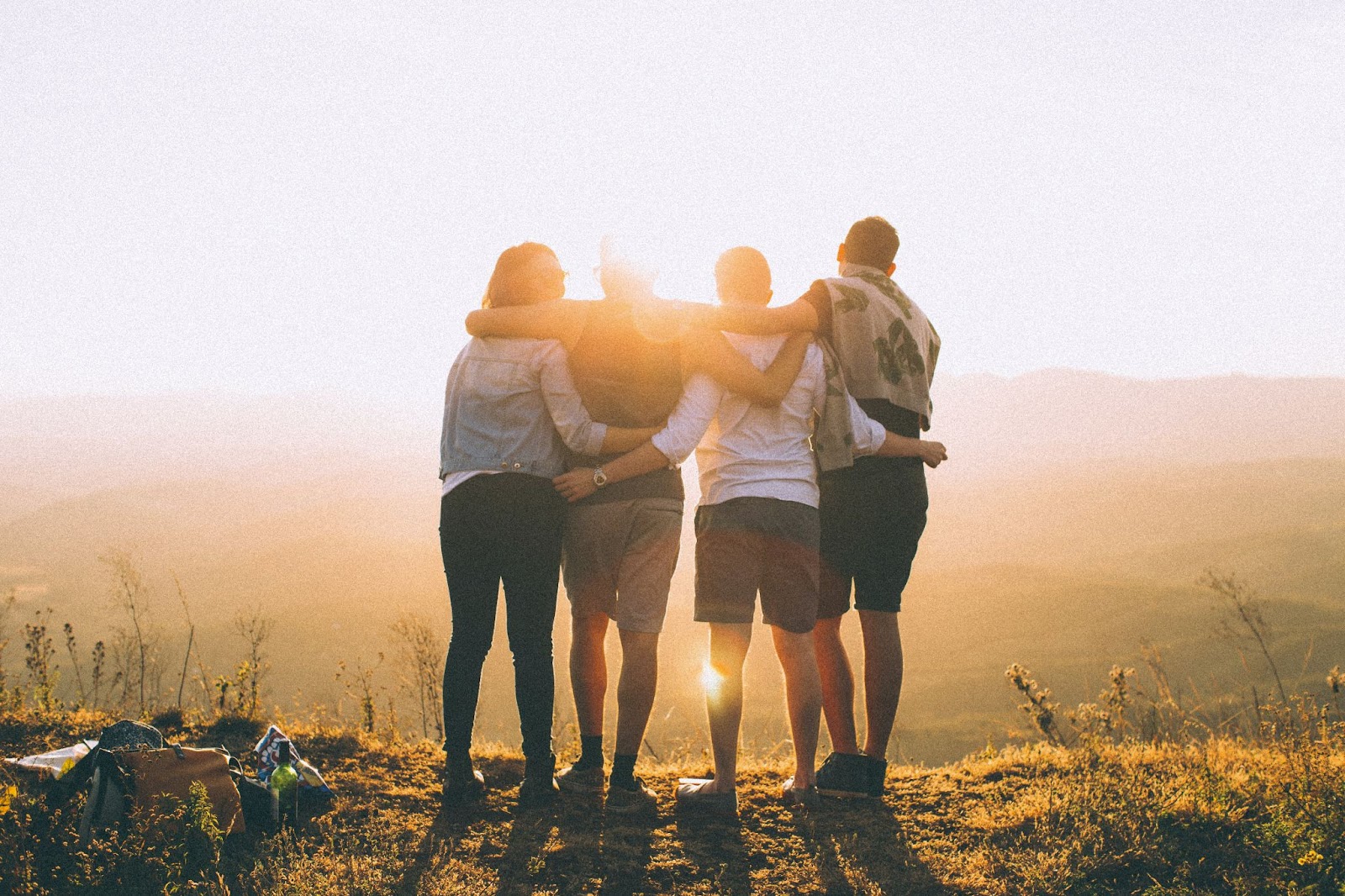 four friends with their arms around one another, watching a sunset with mountains in the background