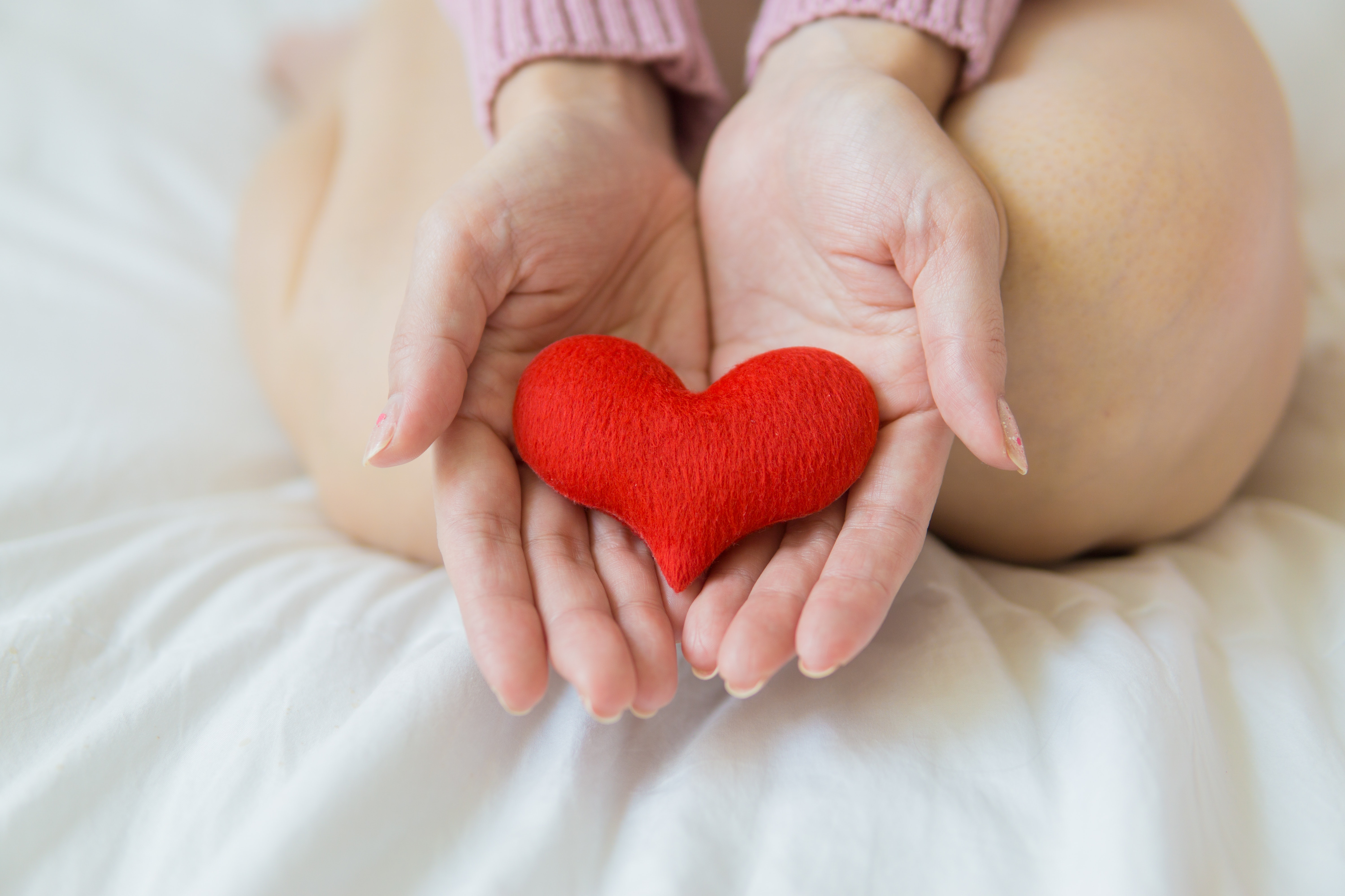 a woman's hands holding a soft read heart plushie. She has her feet curled underneath her and she is sitting on white sheets.