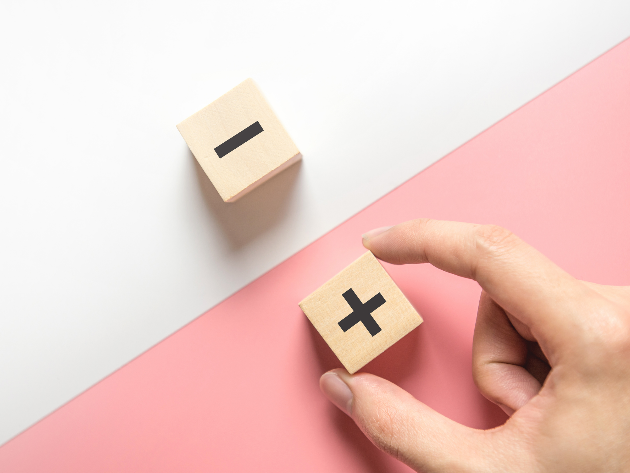 two wooden blocks - one with a minus sign and one with a plus sign - each on top of a table. The plus is on the pink half of the table, the minus on the white.