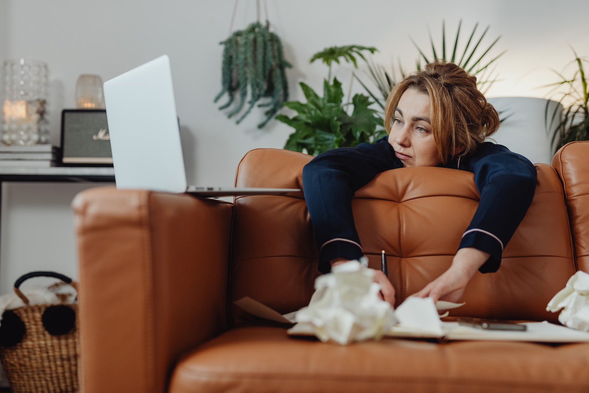 a laptop sits on the arm of a leather chair and a woman is slumped over the back from behind the couch. She's staring at the laptop, stressed or bored. Crumpled papers surround her.