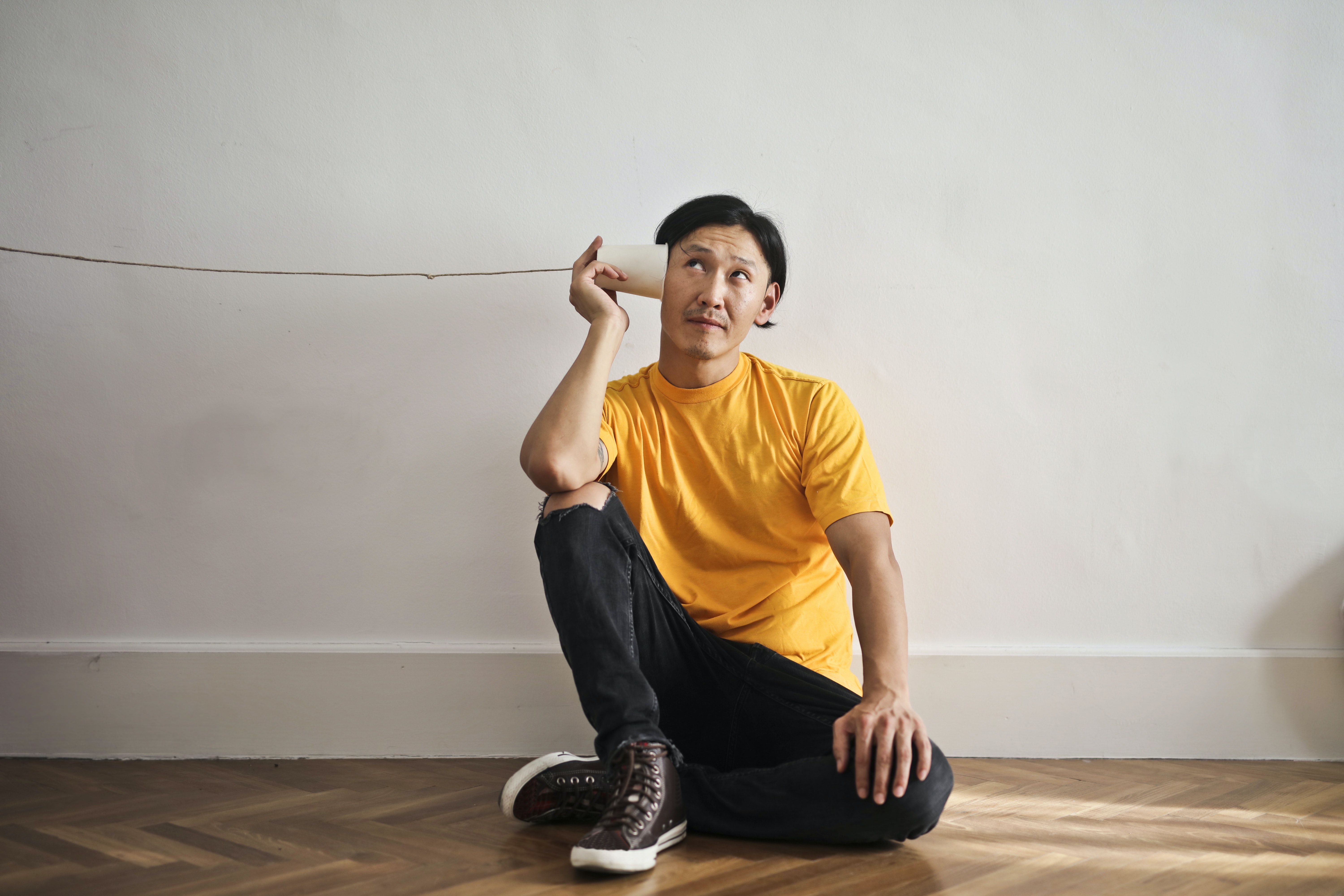 an asian man holding up a paper cup to his ear with a tring coming out at the end. He looks to be listening for something.