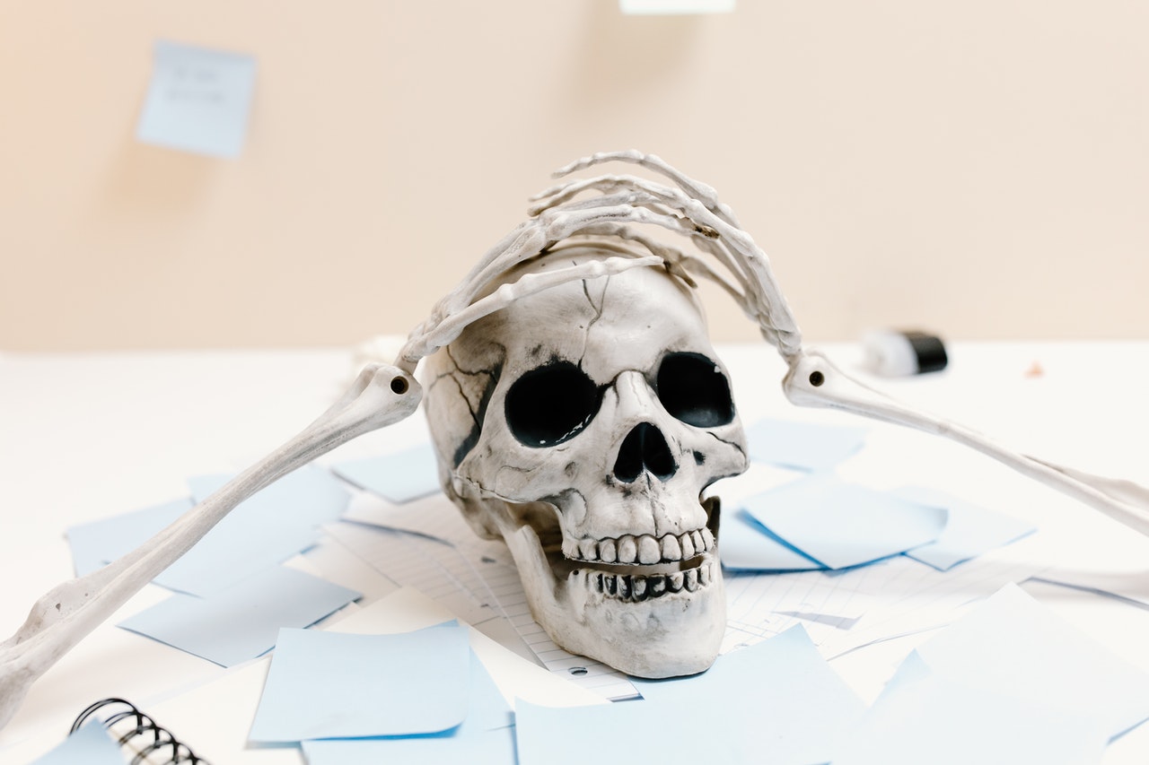 A skull on top of a white desk that is covered with note cards and post-it notes. There are two skeleton arms cupping the skull.