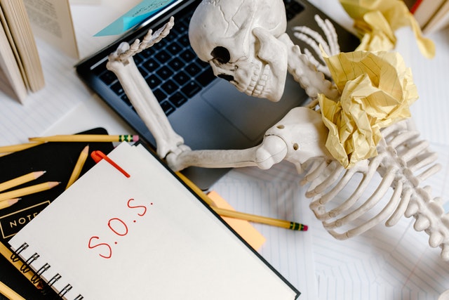A skeleton lying on top of a laptop with crumpled papers surrounding it. "sos" is written in red on a paper next to it