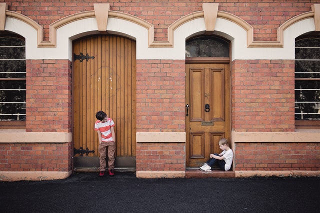 two children sin separate doorways. One child looks lonely and is sitting, while the other is standing with his hands in his pockets and peeking over at the lonely child.