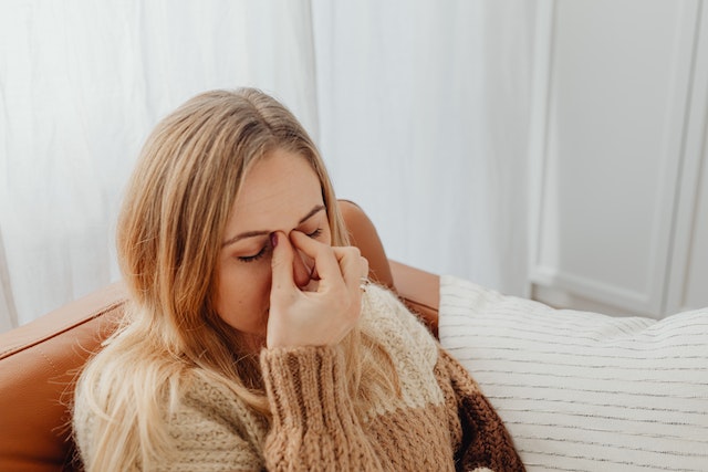 A blonde woman sitting down with her eyes closed and pinching her nose as if she's uncomfortable.