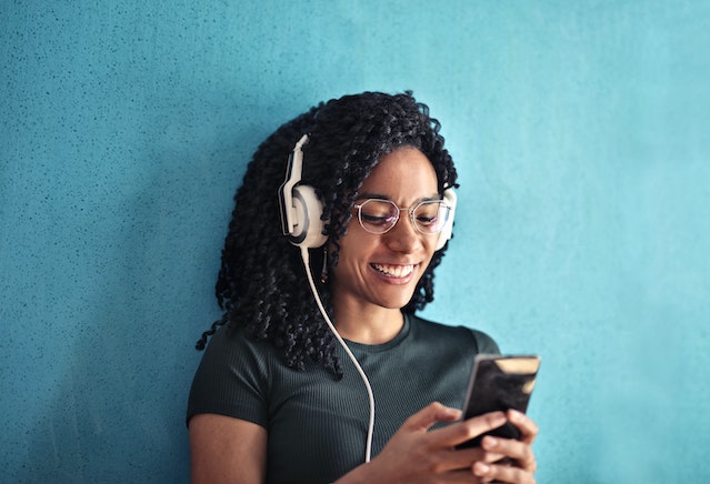 A woman with curly hair standing with her back against a blue wall. She has white headphones on and she is smiling down at her phone.