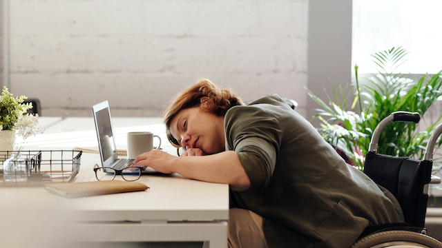 a disabled woman sitting at her desk and wheelchair. she is asleep on her laptop