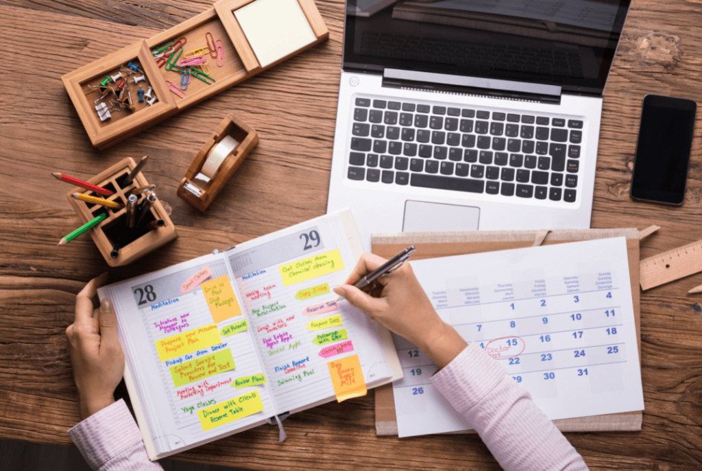 A birds-eye view of a wooden desk with a laptop, stationary (notepads, paperclips, pins), and a monthly calendar and opened daily planner. Two hands are visible, one holding a pen and making notes in the planner. The planner is full of color-coded entries (in yellow, orange, pink, and green) and post-its. It looks very crammed.