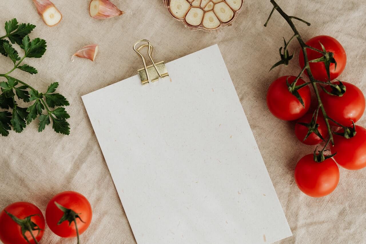 A clipboard with a blank page, surrounded by tomatoes, a garlic bulb, shallots and some parsley stalks.