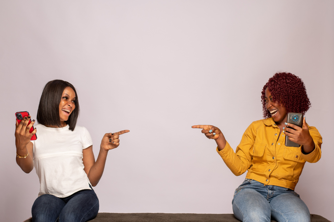 Two Black women sitting on a bench in a waiting room. They both have their phones in their hands and are pointing to each other and smiling and laughing.