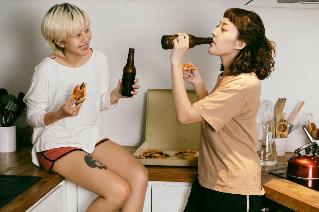 Two friends in a kitchen, one sitting on the countertop, one leaning against it. They're smiling, drinking beer and eating pizza from an open carton.