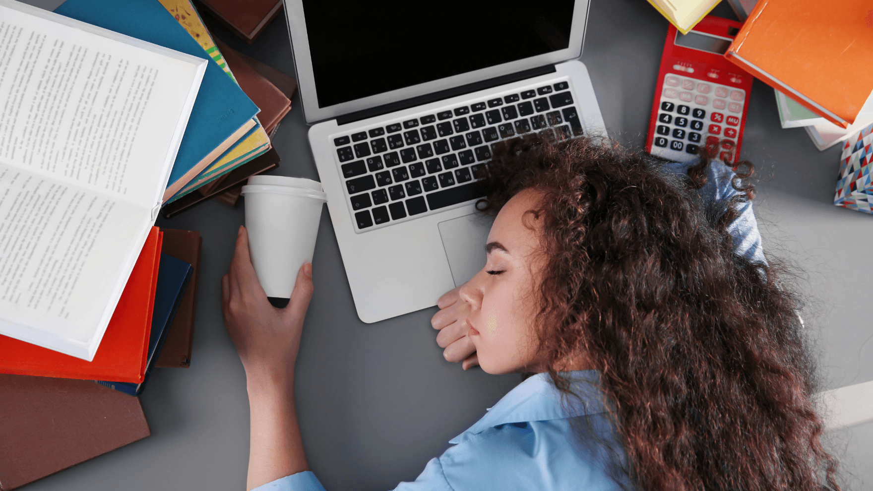 A top-down view of a woman with long curly hair lying on her desk with a laptop open and various books and notebooks scattered about. She's holding a cup of coffee in her hand while she sleeps.