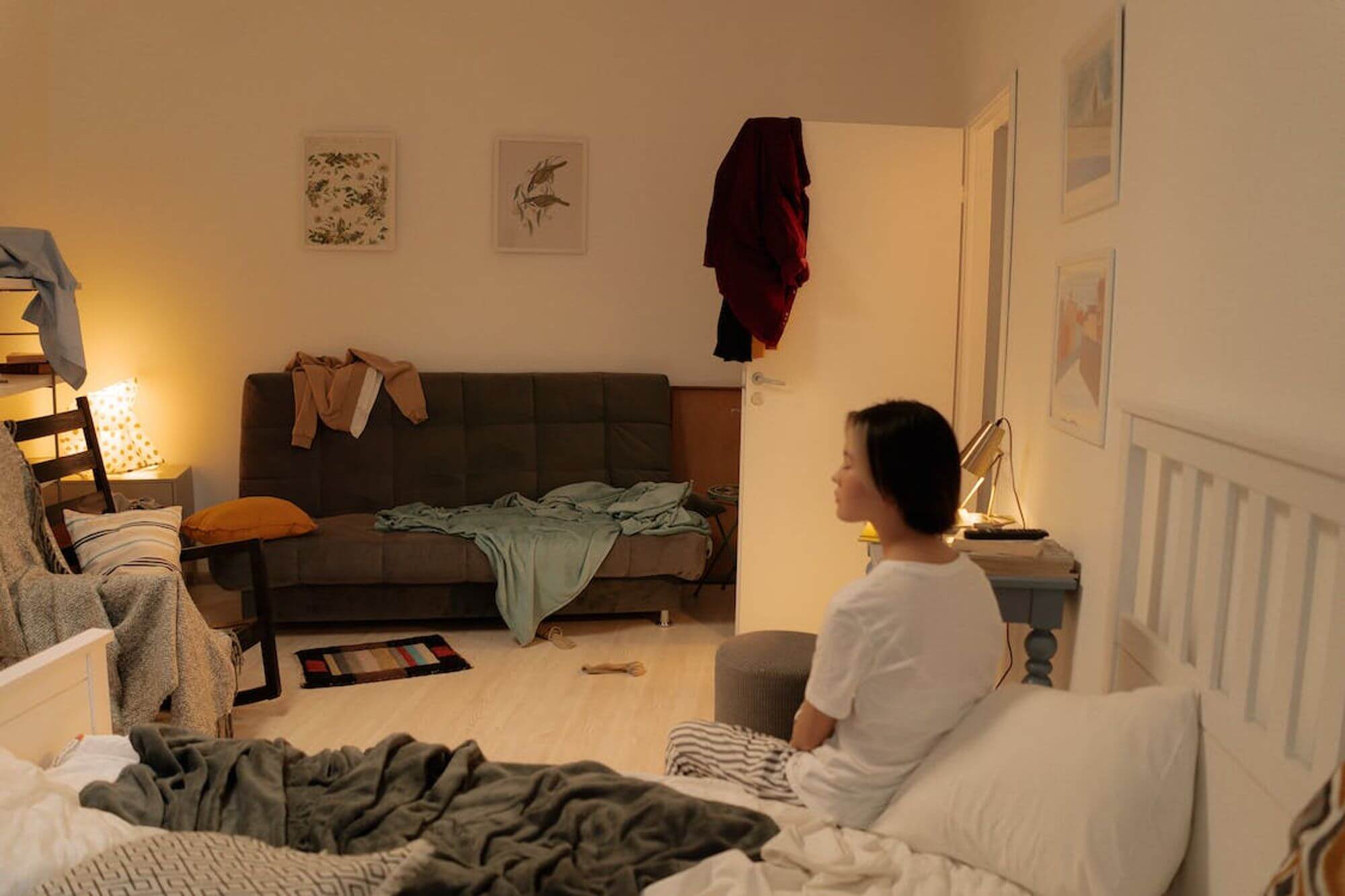 A woman sitting on her bed and looking defeated by the messy house surrounding her.